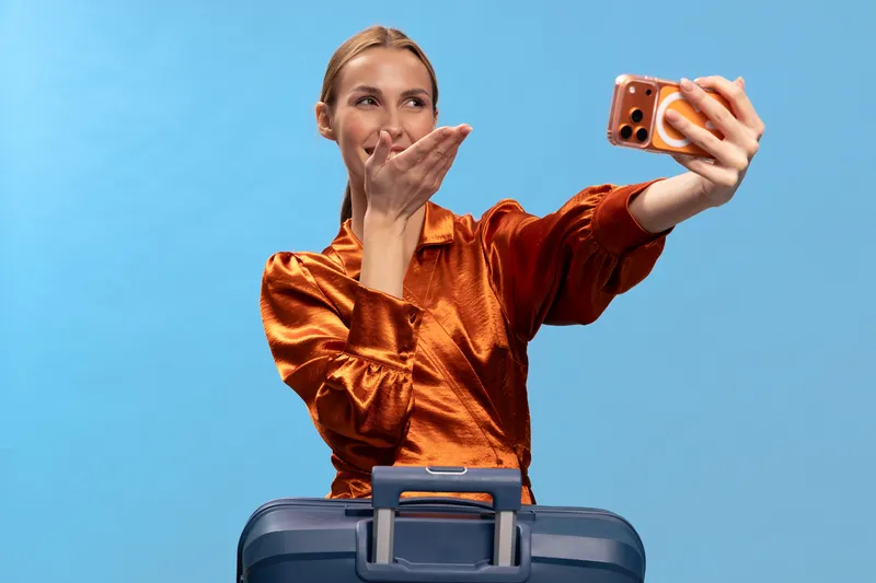 Young woman in shiny orange blouse takes a selfie with smartphone while standing behind suitcase. Blue background. Concept of travel, vacation, and modern communication.