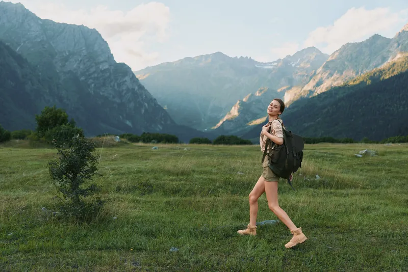 Woman hiking outdoors with a backpack in a meadow, mountains in the background, enjoying sunny weather and natural scenery.
