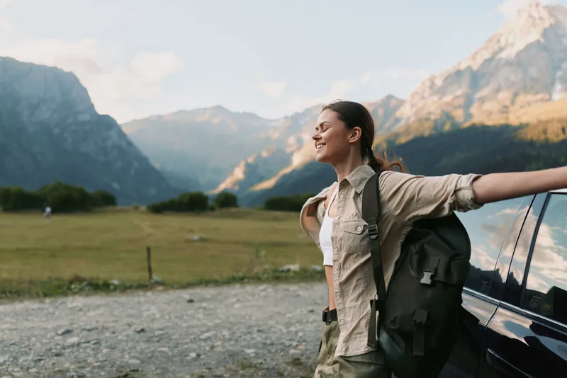 A young woman with a backpack leans against a car in a valley surrounded by towering mountains after a hike, savoring the expansive outdoor scenery.