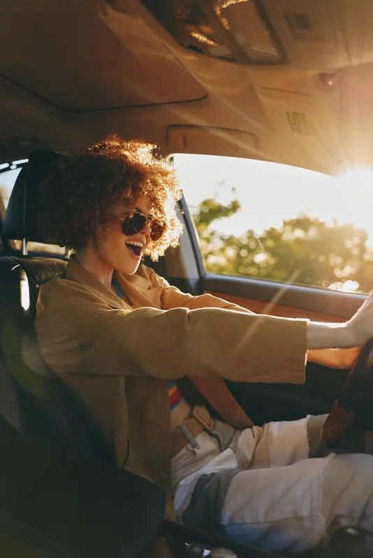Woman driving a car with sunglasses, joyful smile, relaxed casual mood, warm retro film color, sunlit interior, golden hour glow, candid lifestyle portrait isolated on a sunlit road, carefree moment,