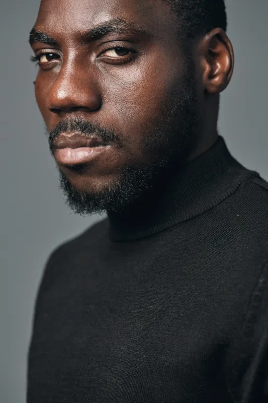 African man portrait with serious expression and beard wearing black turtleneck sweater in creative conceptual visual style with closeup on face and textured skin details.