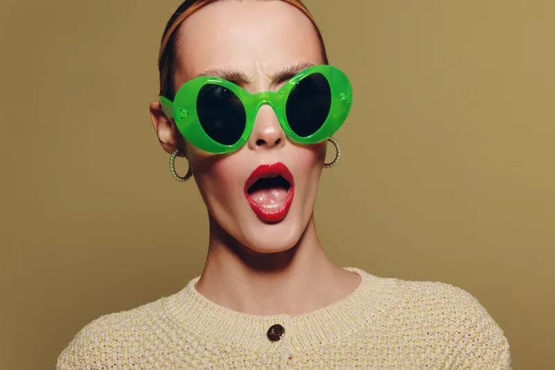 Surprised woman with bright red lipstick wearing large green sunglasses and beige sweater on neutral background. Concept of fashion, style, expression, and modern accessories.