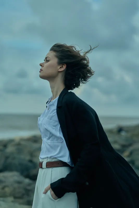Windy coastal scene featuring a woman in a dark coat over a white shirt, belt visible, standing on a rocky shoreline, facing the wind under a moody cloudy sky, motion and mood.