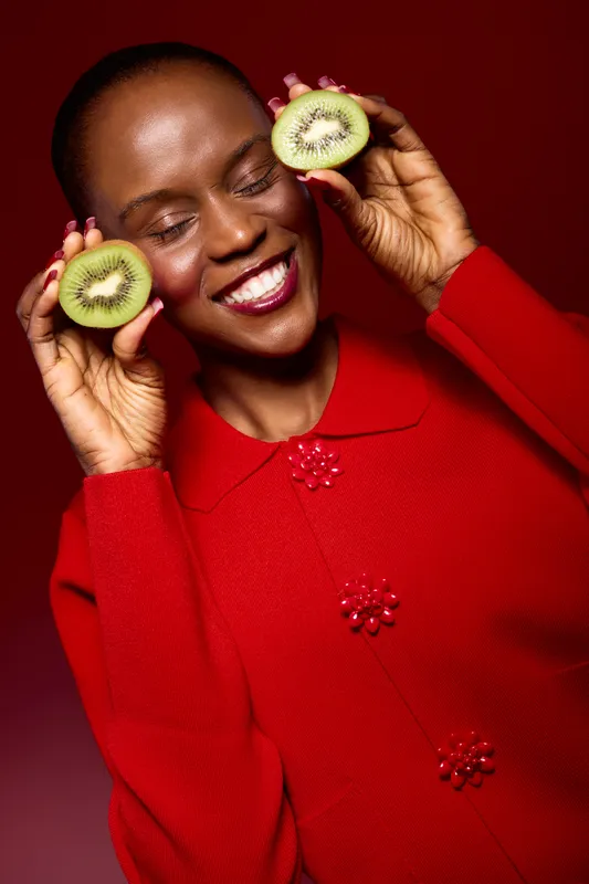 Smiling black woman with dark skin holds two kiwi halves near her face, wearing a red sweater. Studio portrait on red background with fresh fruit, wellness, and joyful mood concept.