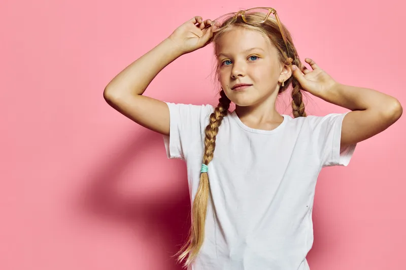 Young girl with braided hair wearing glasses and a white t-shirt poses confidently against a pink background. Studio portrait with soft lighting.