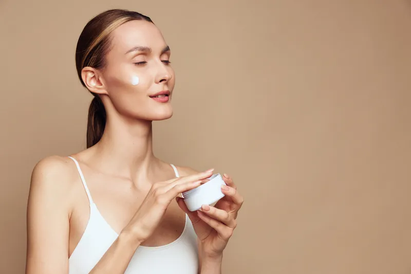 Young woman with brunette hair applies white face cream on her cheek while enjoying a skincare routine against a beige background in soft natural light.