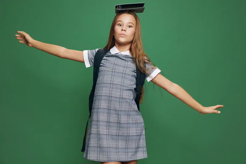 Girl child balancing with arms outstretched against green background, wearing checkered dress and vest in studio. Playful pose of kid with long hair and focused expression.