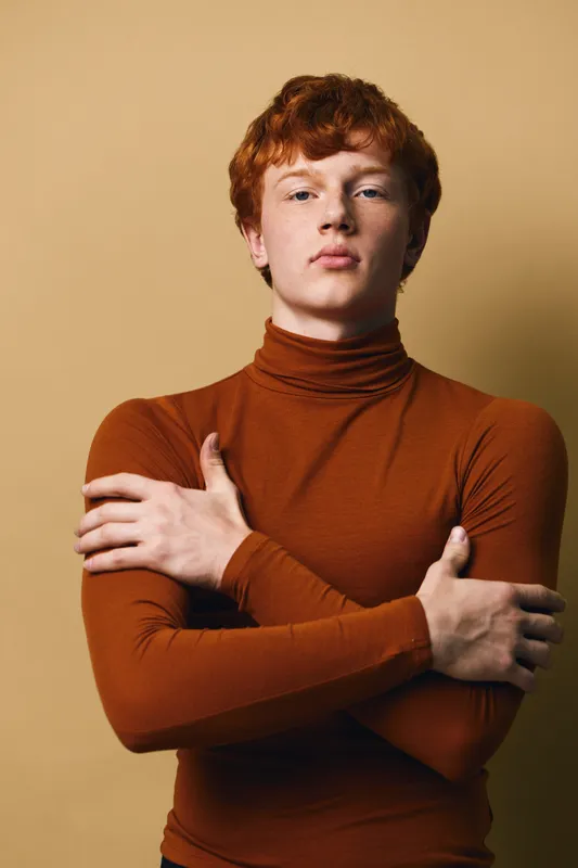 Young man with red hair wearing brown turtleneck sweater stands with crossed arms looking confidently at camera against plain beige background portrait fashion style.