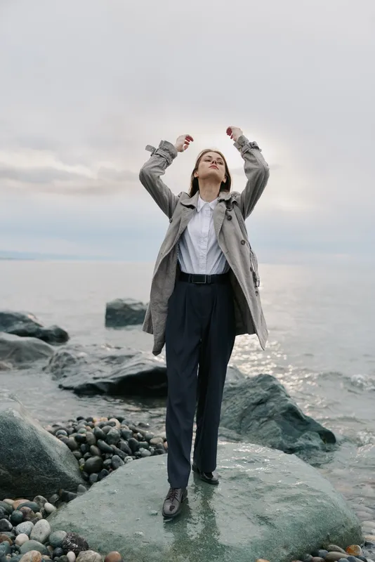 Young woman in coat stands on rocky shore near calm sea under cloudy sky, raising arms. Concept of freedom, nature, fashion, and peaceful outdoor moment by the water.