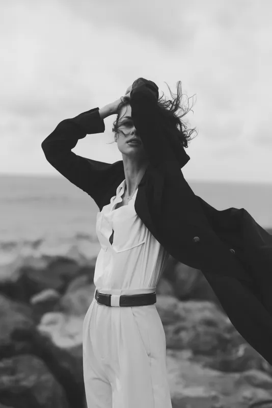 Windy seaside fashion scene with a model in white jumpsuit and flowing dark coat, hair swept by the breeze, capturing a monochrome mood and coastal freedom, sharp composition, textured fabric against