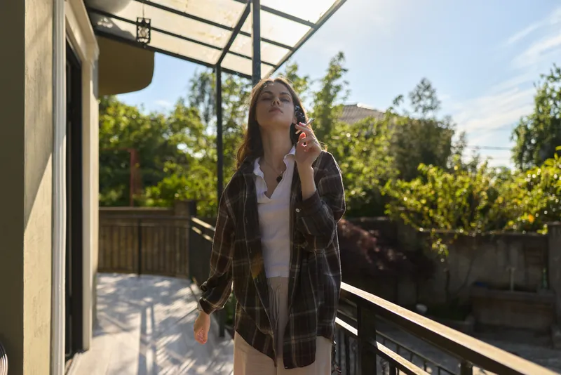 Cheerful woman outdoors on a sunlit balcony, wearing a plaid shirt and light dress, enjoying fresh air, relaxed pose, natural colors, leafy background, casual lifestyle moment