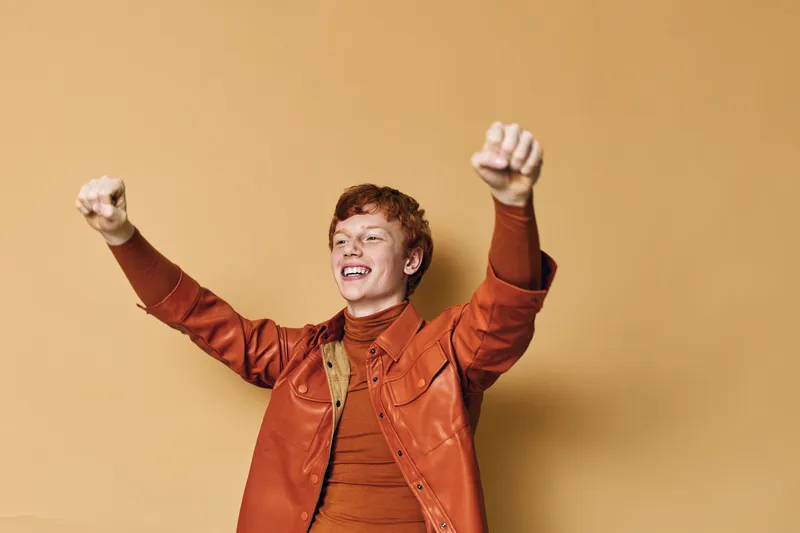 Young man with red hair wearing orange jacket and turtleneck smiling and raising fists in excitement against light brown background. Celebration happiness and success expression.