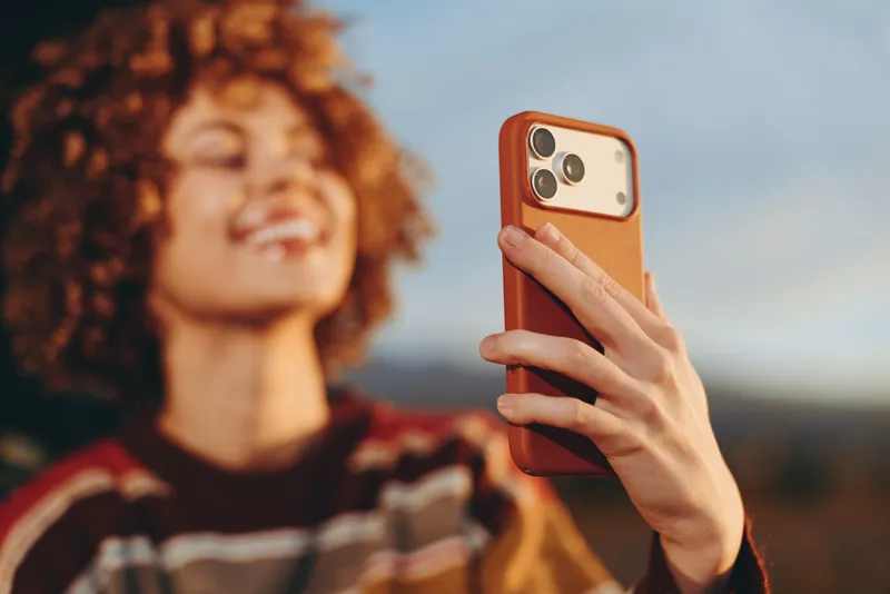 Woman lifestyle outdoors, curly haired woman smiling while holding smartphone for a selfie, warm golden sunlight, shallow depth of field, soft sky and blurred landscape background