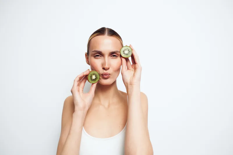 Young woman with clear skin holding two halves of kiwi fruit near her face, puckering lips, minimal makeup, white background, fresh and healthy concept