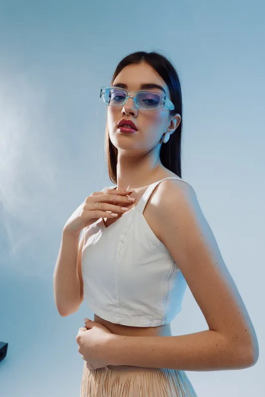 Young woman in white top and transparent sunglasses poses confidently against a blue background. Concept of beauty, fashion, and modern style in studio lighting.