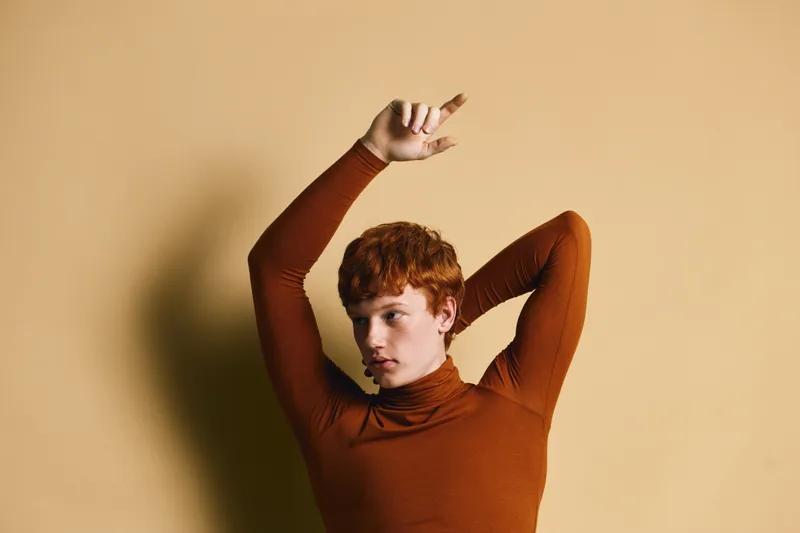 Portrait of young man with red hair and brown turtleneck posing with arms raised against beige background in studio for fashion concept.