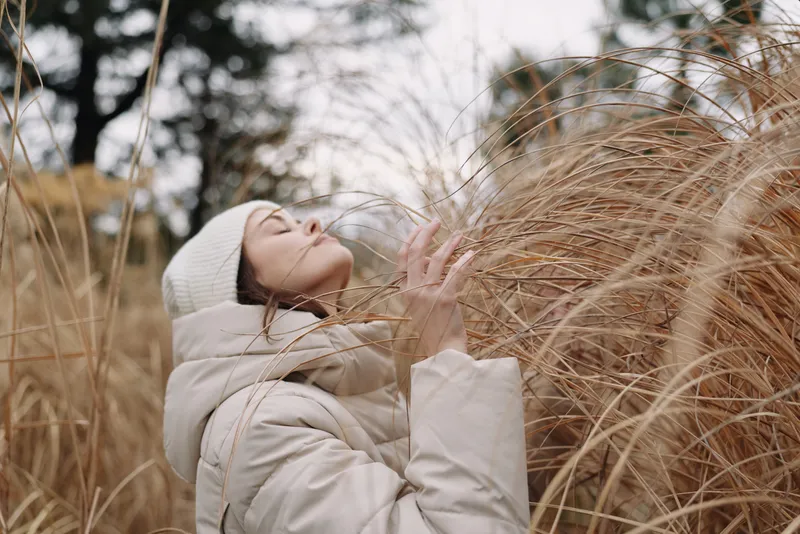Windy outdoor portrait of a woman in a light coat amid tall grasses, calm pose, soft sunlight, autumn atmosphere, cozy winter fashion and natural movement in nature, serene mood.