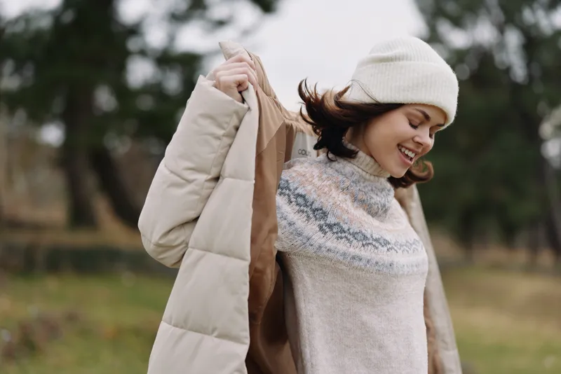 Woman outdoors in a cozy knit sweater and beanie, smiling as she holds a warm coat over her shoulder in a park setting with trees, autumn light, and relaxed fashion vibe.