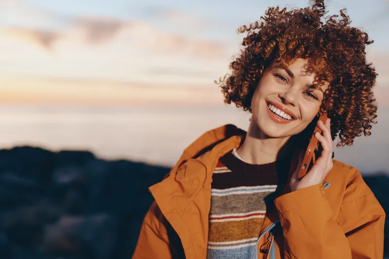 Woman with curly hair smiling outdoors at sunset, wearing orange jacket, holding phone near ear, warm lifestyle portrait with soft breeze and golden light on blurred landscape distant