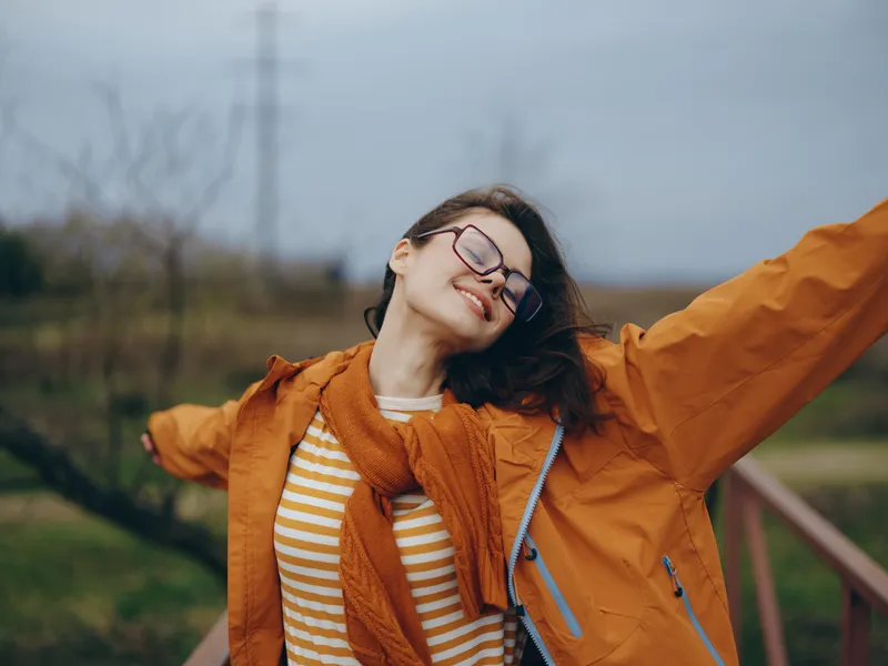 Woman lifestyle outdoors, orange jacket, glasses, smiling and wearing headphones, enjoying fresh air near park trail, cloudy sky background, casual candid portrait, soft natural light