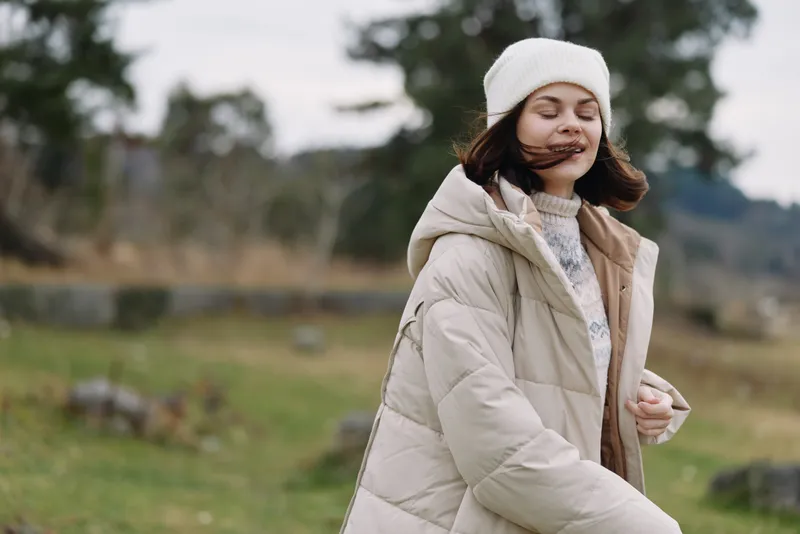 Girl outdoor winter coat walks through windy countryside, smiling softly in a white beanie, cozy mood and tranquil natural landscape.