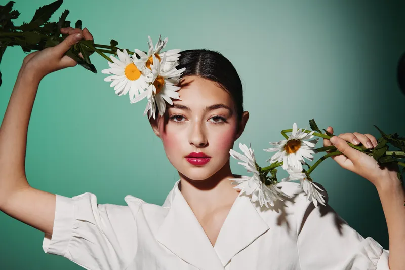 Young woman with dark hair styled holding white daisies over her eyes wearing a white blouse. Studio portrait on teal background. Fashion concept with beauty contrast and natural elements.