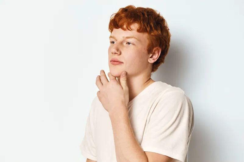 Young man with red hair and light skin standing on white background, wearing beige t shirt looking away with thoughtful expression in studio portrait