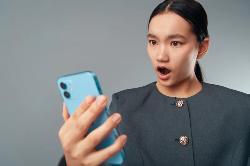 Woman with surprised expression holding smartphone looking at screen in gray background, technology, communication, emotion and reaction for digital device usage and social media.