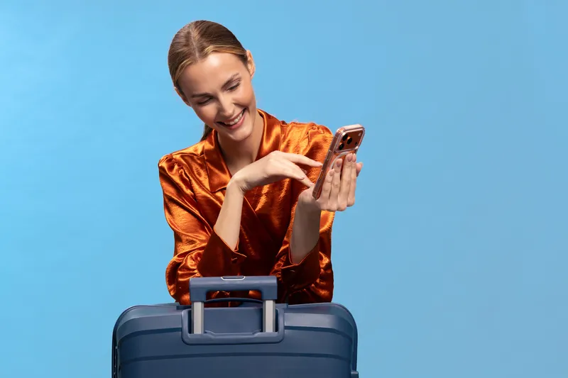 Smiling woman in orange blouse stands behind blue suitcase, looking at smartphone with excitement on blue background. Concept of travel, technology, happiness, and modern lifestyle.