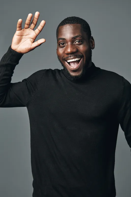 African man with dark skin waving hand and smiling in creative visual designer style. Portrait of happy black male in black sweater on gray background with friendly gesture and expression.