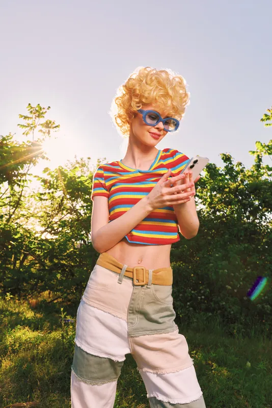 Young woman with a blonde curly wig, striped crop top and patchwork pants, posing outdoors in bright sun, retro vibe, casual lifestyle, vibrant colors, playful mood, summer atmosphere.