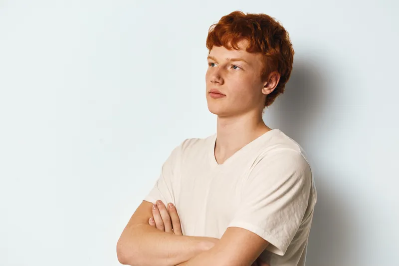 Young man with red hair standing with arms crossed wearing white t shirt looking aside isolated on white background with thoughtful expression and casual pose.