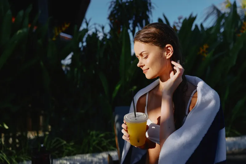 Cheerful woman outdoors, relaxing with a cold beverage, sunlit skin, soft breeze, towel draped, lush tropical plants in the background, natural colors, casual lifestyle and sunshine