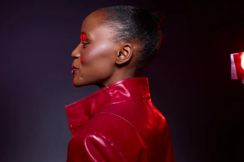 Profile of a dark skinned African woman wearing a red leather jacket with glossy makeup and slicked back hair. Studio portrait with dramatic lighting and a dark purple background.