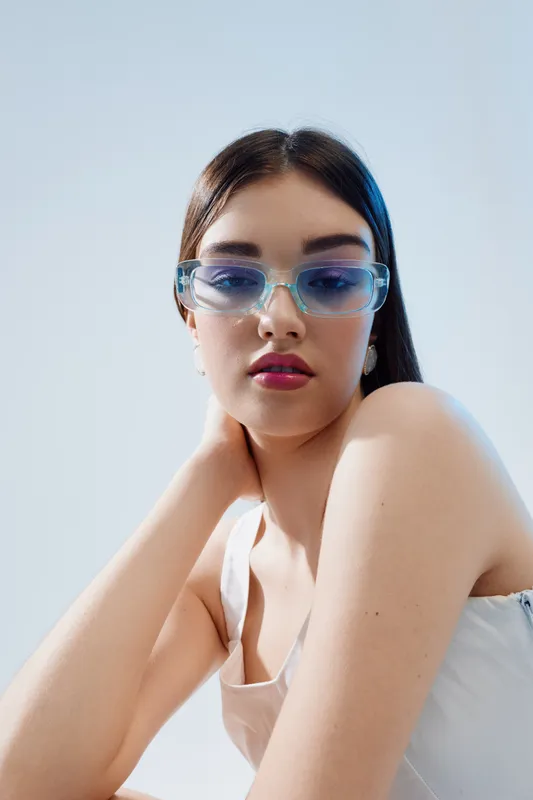 Young woman with long dark hair wearing transparent blue sunglasses poses against a light background. Fashion and beauty concept with minimal makeup and casual style.