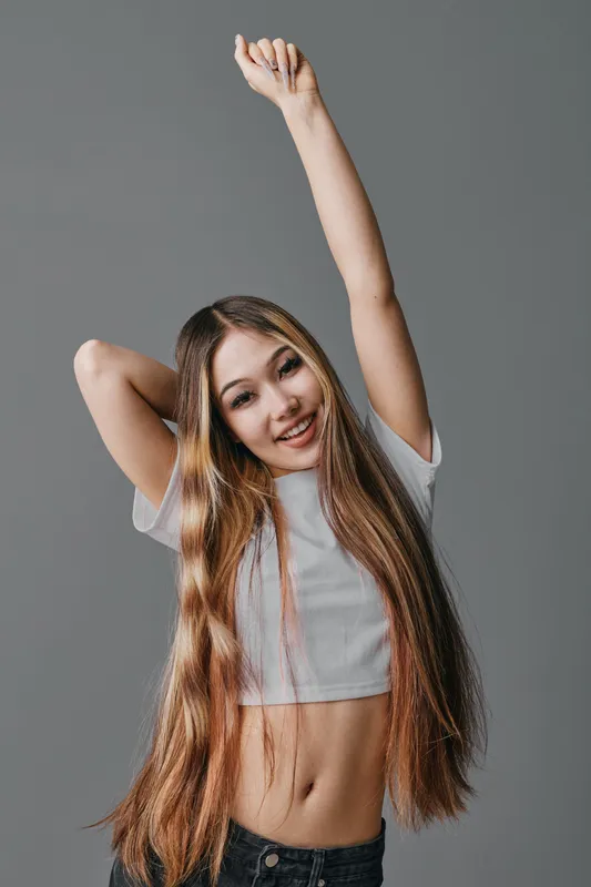Young woman with long hair wearing white crop top and black jeans stretching arm up while smiling on grey background standing in studio space.