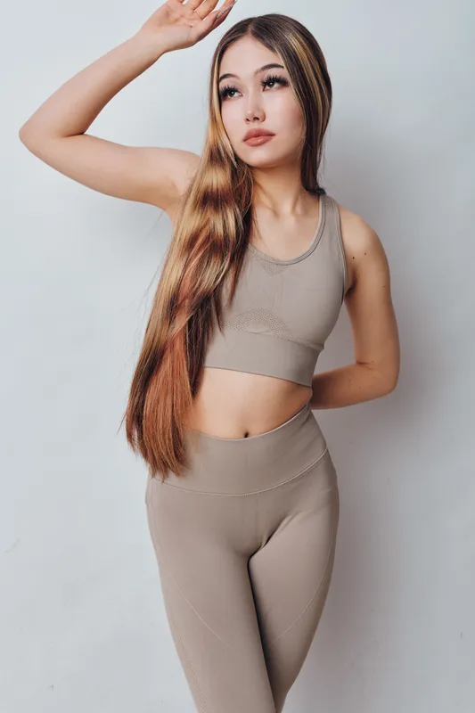 Woman with long hair posing in beige sportswear standing against white background with hand raised above head. Fitness clothing and casual look for active lifestyle and youth.