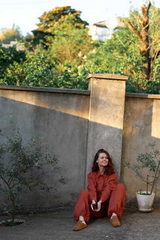 A young woman wearing an orange jumpsuit sits on the ground beside a concrete wall in a sunlit outdoor garden, creating a calm street style portrait