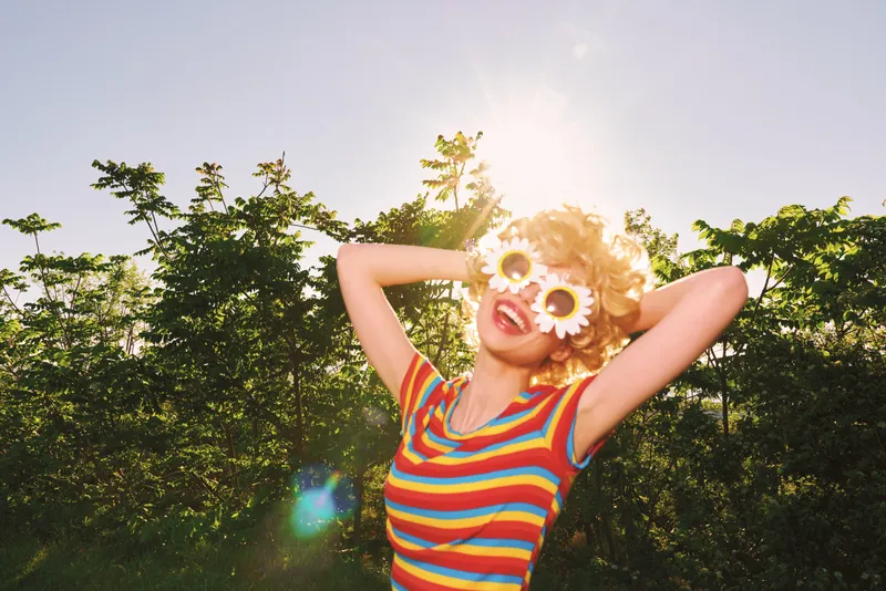Cheerful woman outdoors, wearing a striped colorful shirt and flower sunglasses, sunlit moment, relaxed pose, natural colors, lush greenery background, vintage vibe