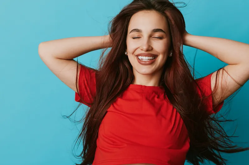 Young woman with long hair and closed eyes smiling happily in red shirt against blue background. Relaxed female enjoying moment with casual pose and natural beauty indoors.