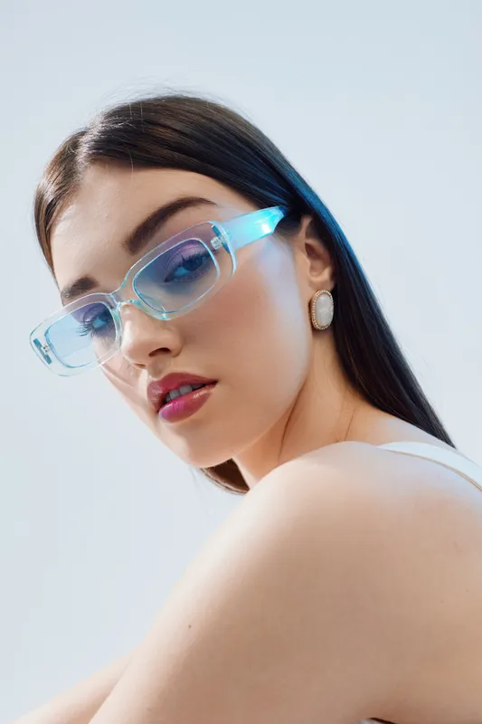 Young woman with long dark hair wearing transparent blue sunglasses and white earrings poses against a light background. Concept of beauty, fashion, and style.