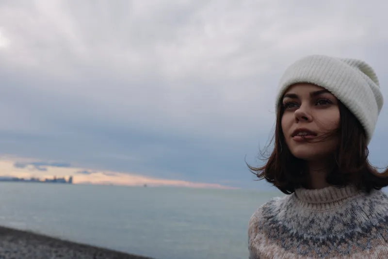 Woman on a beach wearing a knitted sweater and white beanie gazes at the sea along a moody shoreline, capturing outdoor calm and coastal wind at dusk