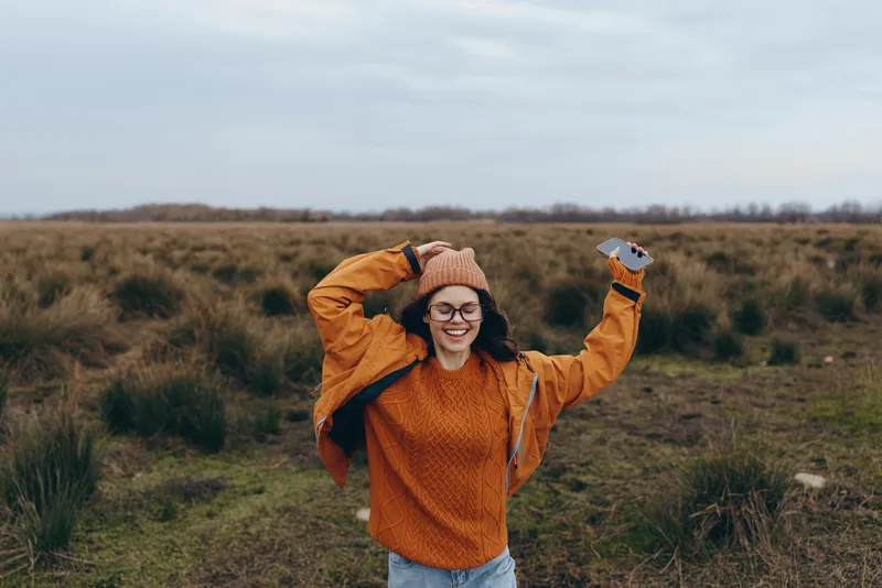 Woman lifestyle outdoors in a countryside field, wearing orange sweater and glasses, smiling with hands behind head under cloudy autumn sky, gentle wind, full body candid portrait.