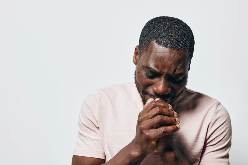 Black man with dark skin praying with closed eyes and hands clasped in front of white background. Spiritual, hope and faith concept with emotional expression and casual clothing.