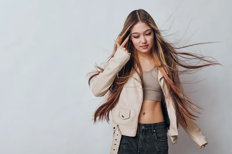 Young woman with long hair wearing beige jacket and crop top standing against gray background with casual style and natural look for fashion and beauty concept.