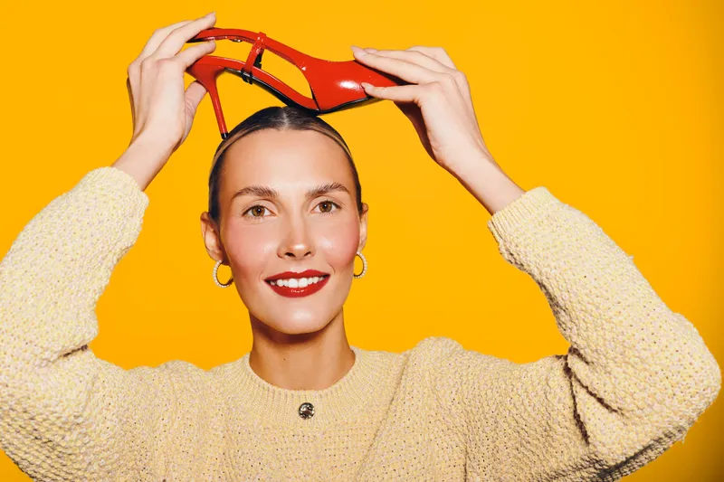 Smiling woman in beige sweater holds red high heel shoes above head on bright yellow background. Concept of fashion, style, beauty, and playful mood in studio lighting.
