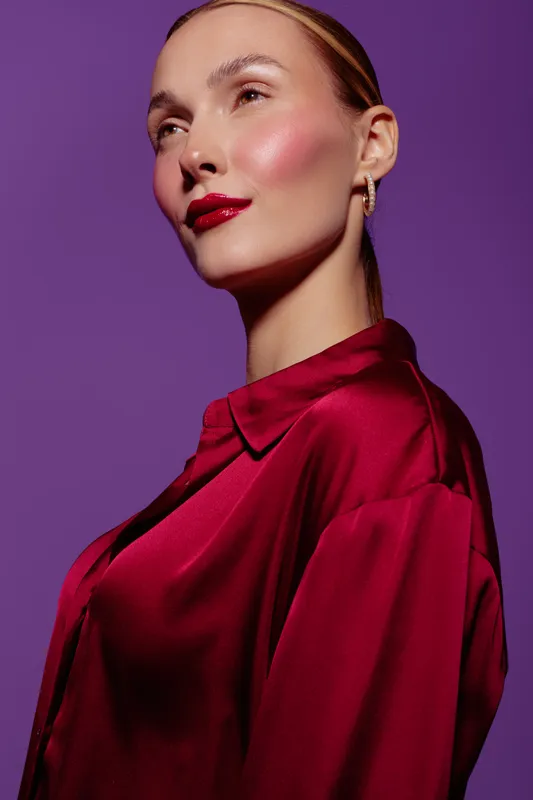 Elegant woman with slicked back hair wears shiny red blouse and bold red lipstick. She poses confidently against a purple background. Concept of fashion, beauty, and style.