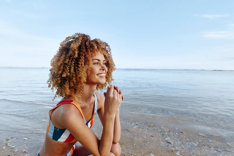 Woman curly hair beach sea seaside smiling sunlit summer moment, sitting on sand at the shoreline, gentle breeze, sunlit skin tone, calm ocean waves, natural colors, soft smile relaxed gaze