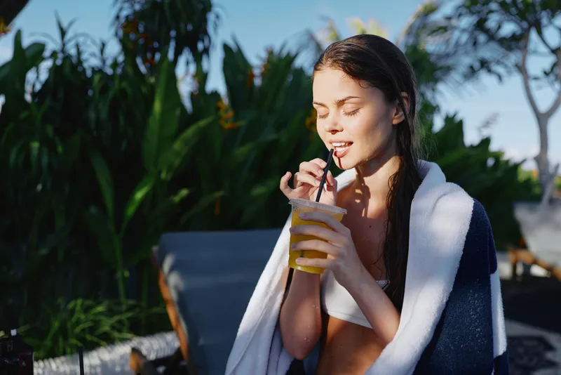 Cheerful woman outdoors, sipping a cold drink through a straw, sunlit moment, relaxed posture, towel draped over shoulders, lush greenery backdrop, casual summer lifestyle
