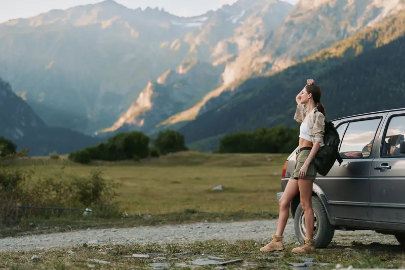 Outdoor scene with a woman standing beside a backpack, leaning on a car, gazing toward distant mountains, sunlit valley, casual travel mood, relaxed and curious and peaceful vibe